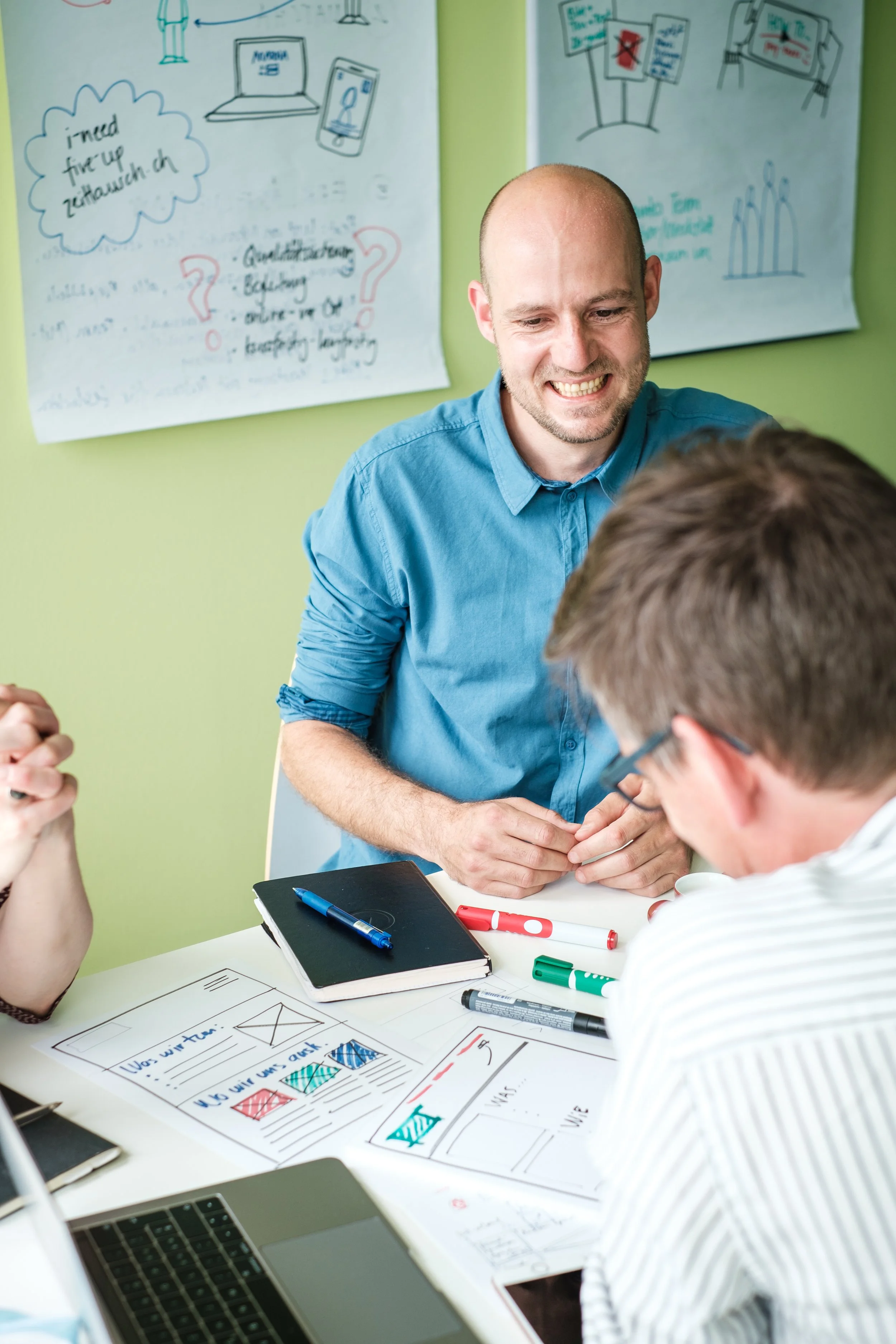 Ein Mann im blauen Hemd, der bei einer Besprechung lächelnd mit einem jüngeren Mann spricht, während sie an einem Tisch mit Diagrammen und Notizbüchern sitzen, im Hintergrund Whiteboards mit Skizzen und Notizen.