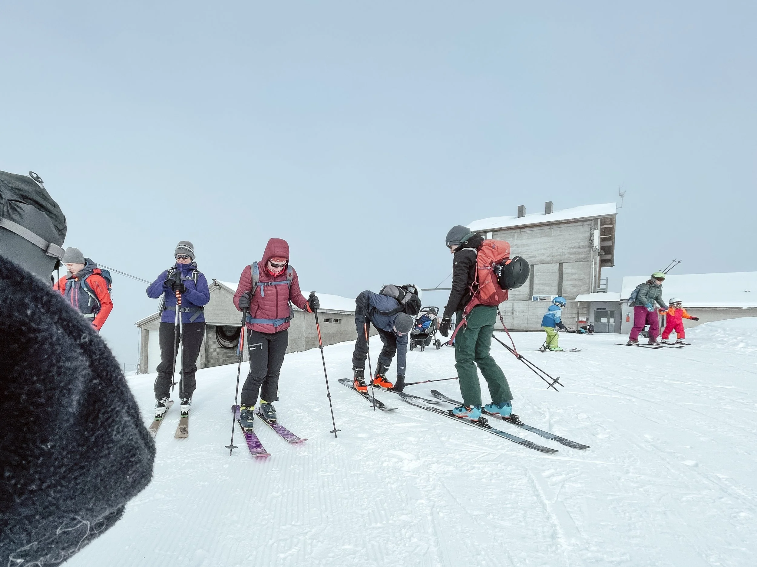 Menschen beim Skifahren auf einer verschneiten Pisten in einer Winterlandschaft, einige bereiten sich vor, andere fahren ab, im Hintergrund eine Hütte.