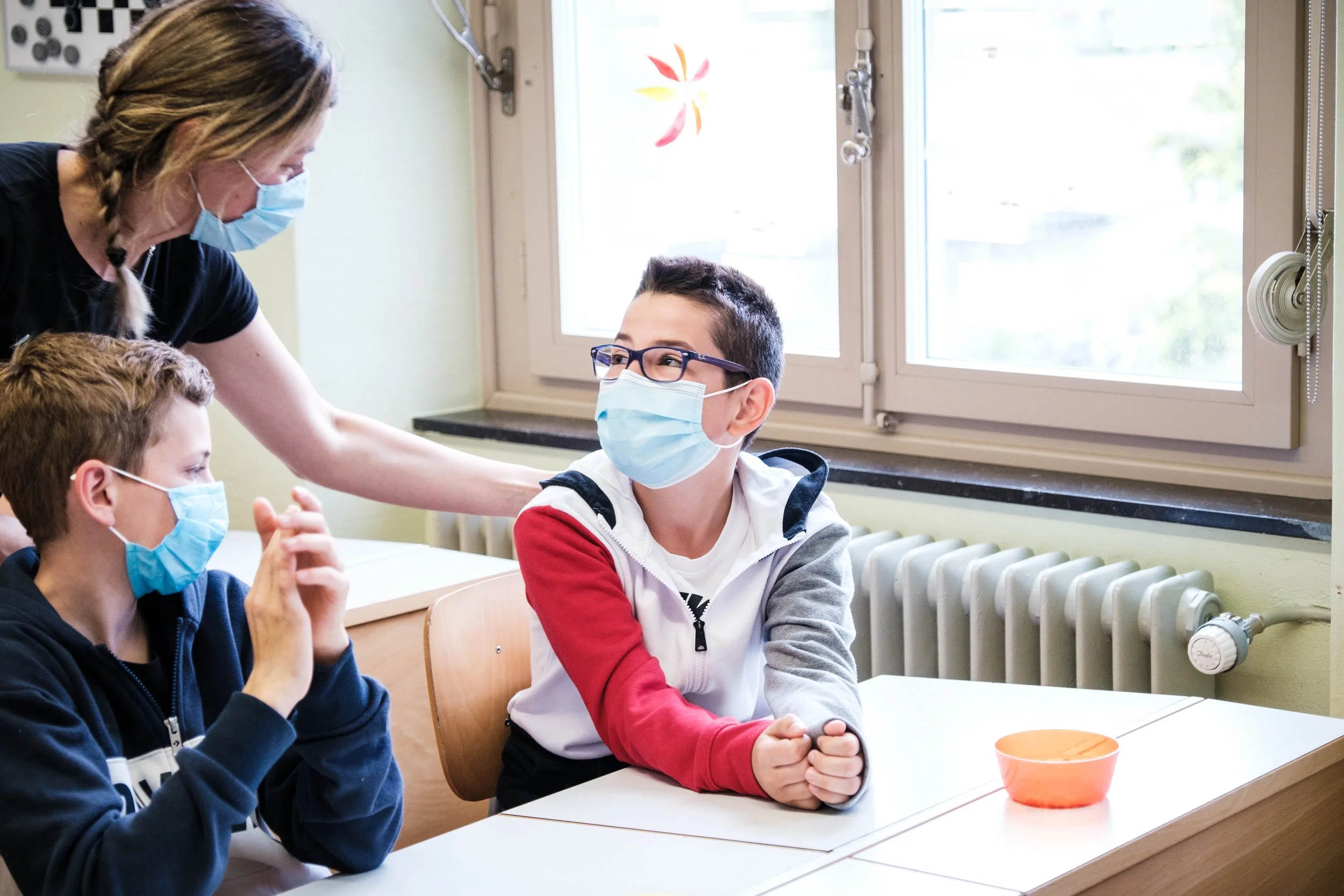 Lehrerin spricht mit zwei Schülern im Klassenzimmer, alle tragen Masken, im Hintergrund Fenster und Heizkörper.