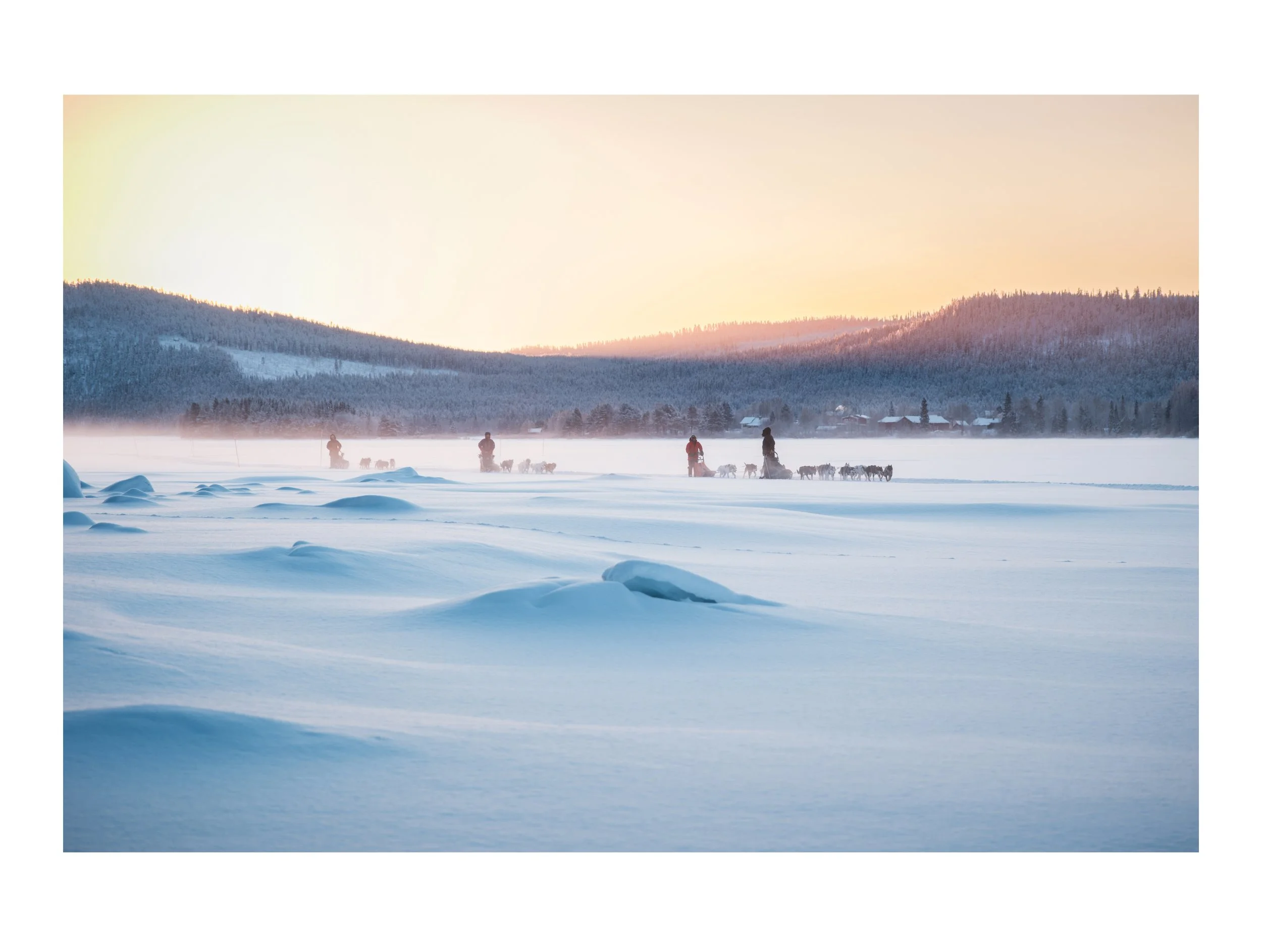 Menschen, Hunde und Hundeschlitten auf schneebedecktem See bei Sonnenaufgang, mit Bergen im Hintergrund.