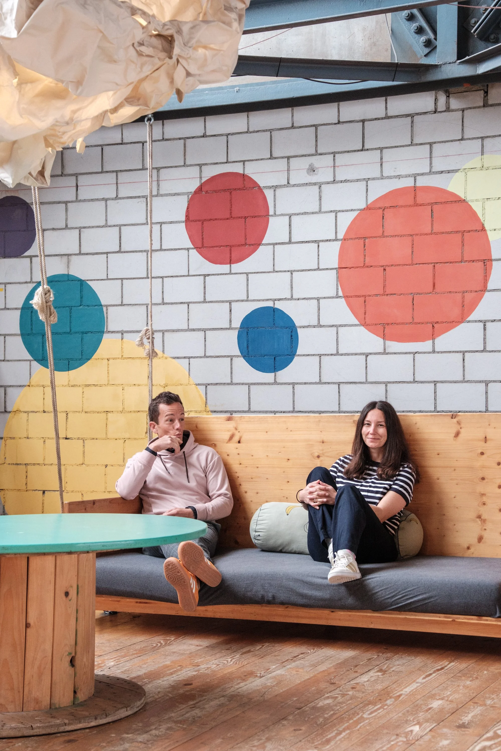 Ein Mann und eine Frau sitzen auf einer Couch in einem Raum mit bunten, großen Kreisen auf der Wand. Der Mann trägt einen hellen Hoodie und die Frau ein gestreiftes T-Shirt. Es sieht freundlich und entspannt aus.