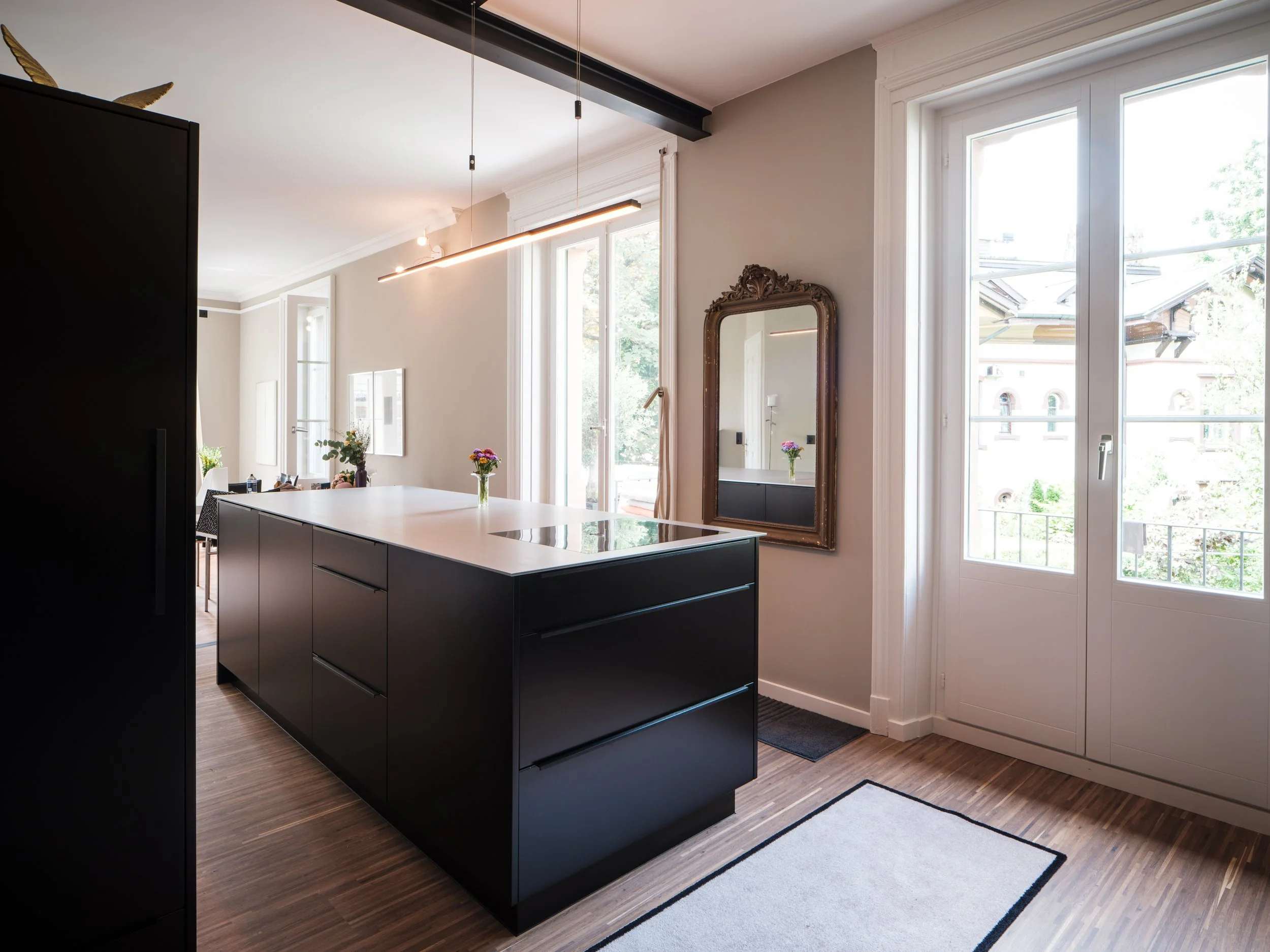 Moderne, offene Küche mit schwarzer Insel, großen Fenstern, einem alten Spiegel an der Wand und einer weißen Tür, die auf einen Balkon führt.