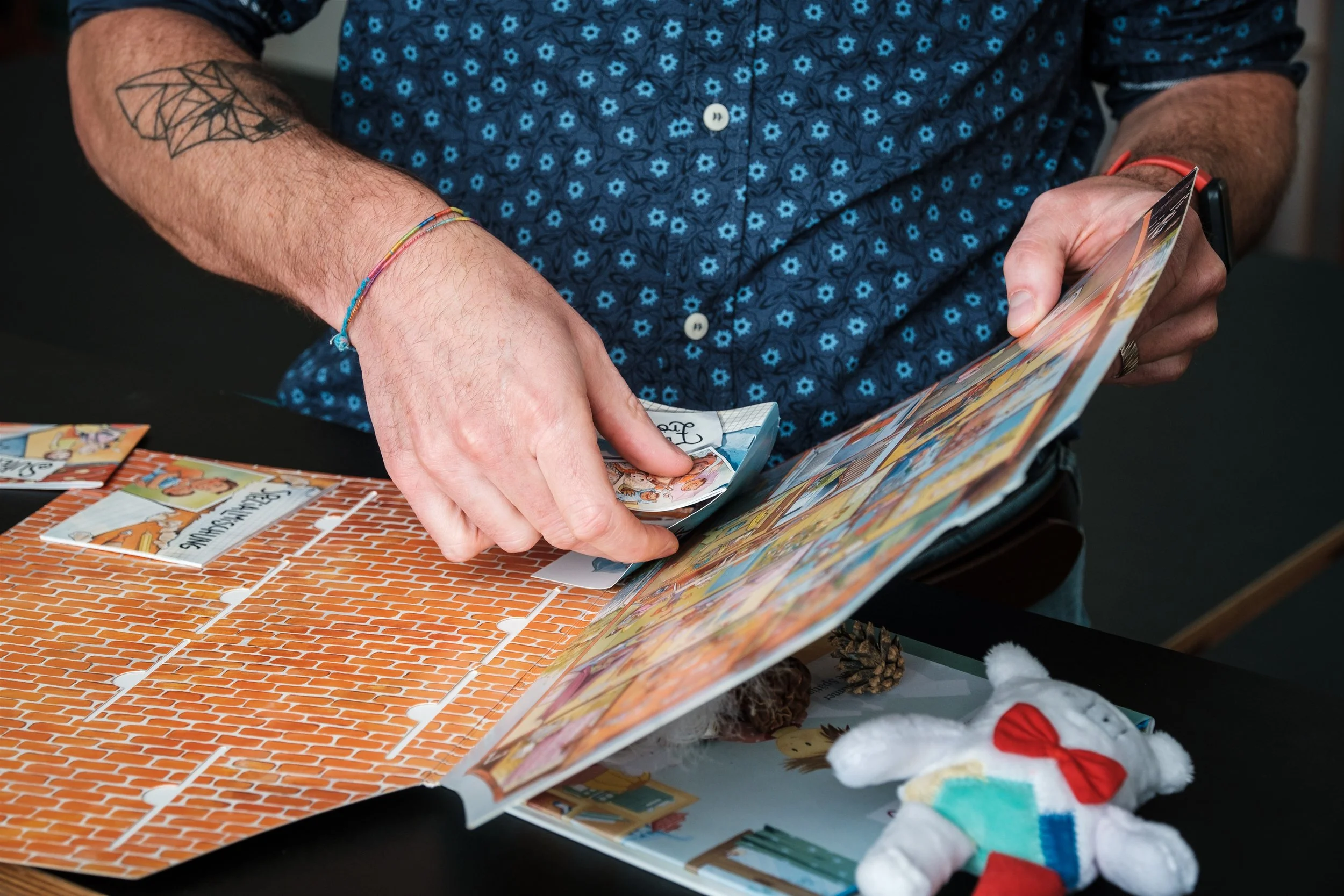 Nahaufnahme einer Person, die ein bunt illustriertes Buch durchblättert, auf einem Tisch mit ausgeschnittenen Comicbildern, einem kleinen Teddybär und Dekoelementen.