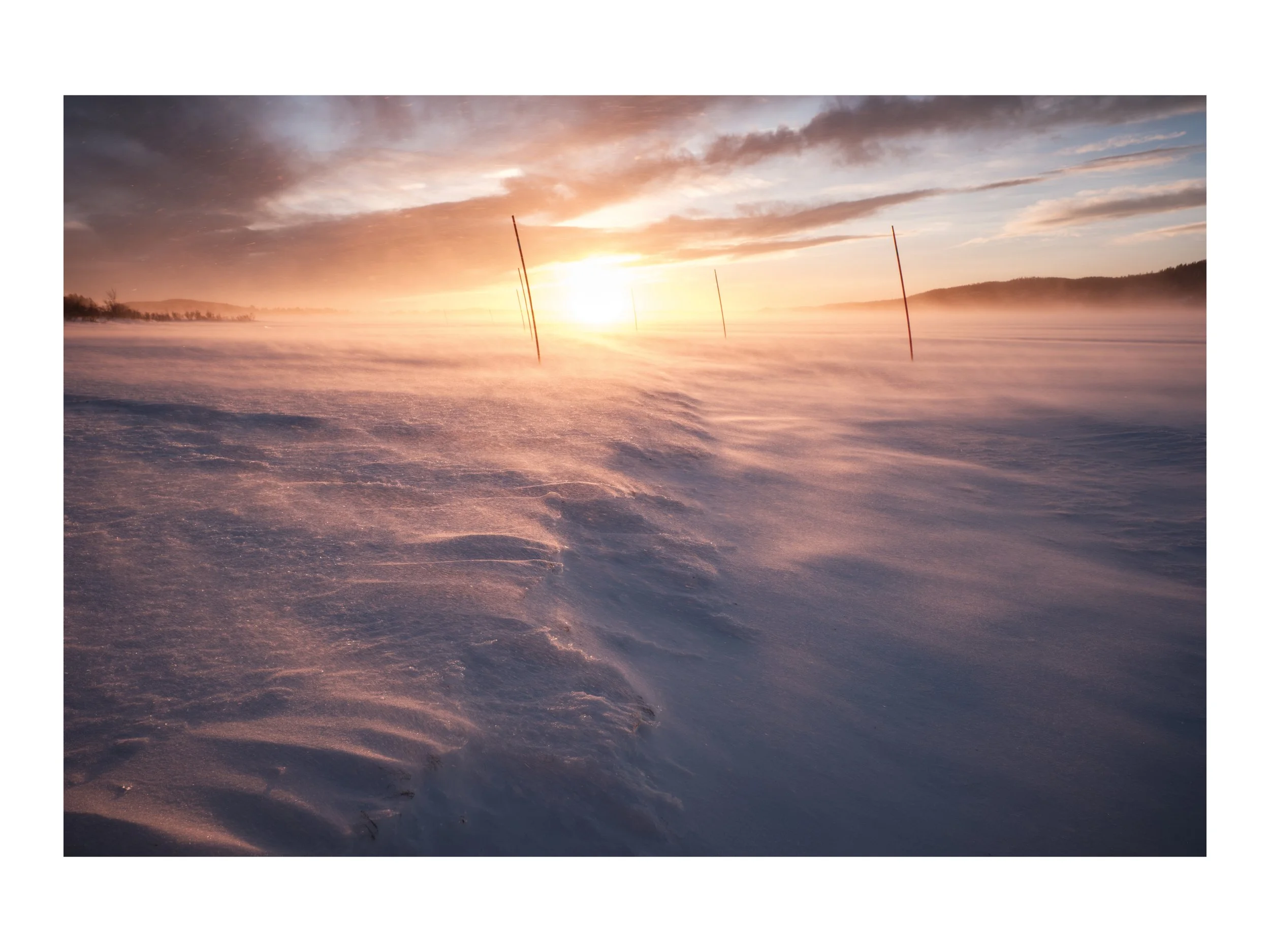 Schneebedecktes Feld bei Sonnenaufgang mit Nebel und Windmühlenstangen im Hintergrund.