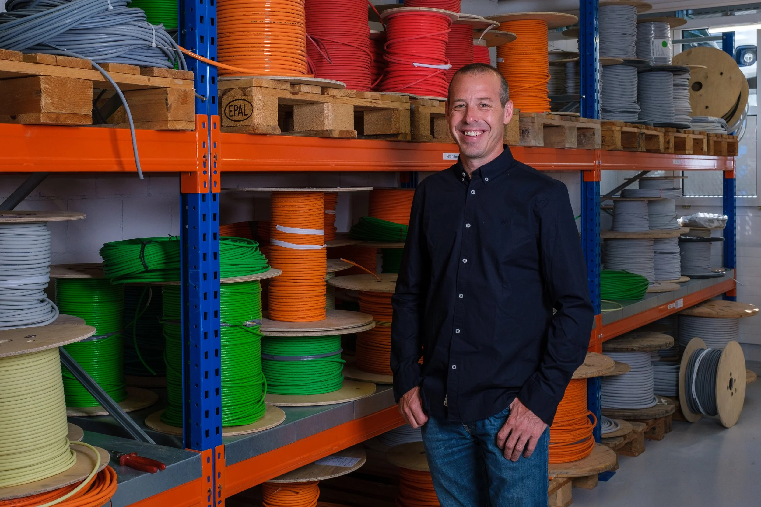 Ein Mann steht in einem Lagerraum mit Regalen, die mit bunten Kabelrollen in verschiedenen Farben wie Orange, Grün und Grau gefüllt sind.