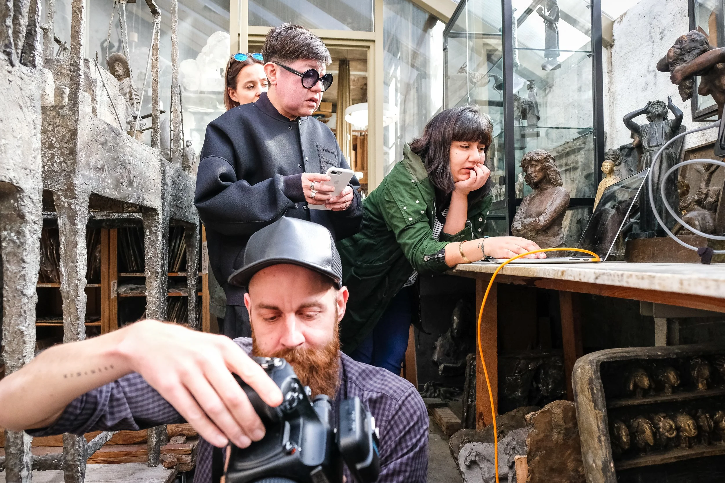 Menschen in einem Museum, mit Keramiken und Skulpturen im Hintergrund, eine Person fotografiert, eine andere betrachtet Objekte auf einem Laptop, eine weitere schaut nachdenklich.