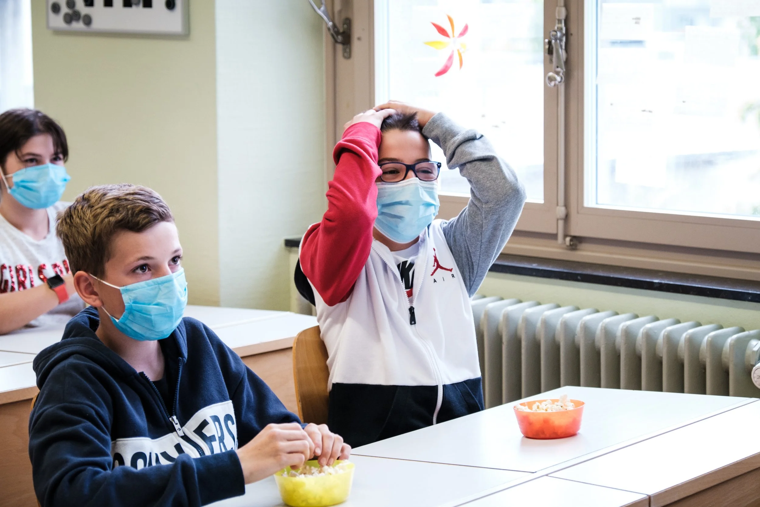 Drei Kinder sitzen in einem Klassenzimmer, tragen Masken und essen Snacks, während eines die Hände auf den Kopf legt.